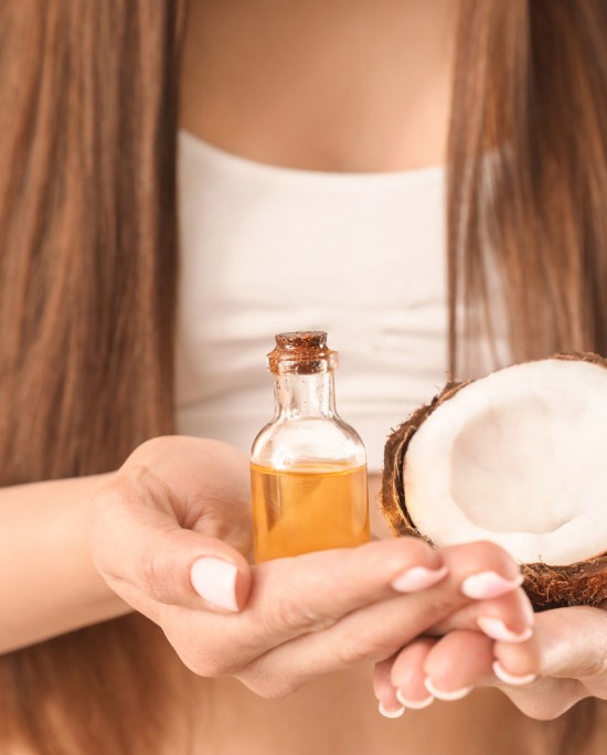 Hands holding a small bottle of coconut oil in front of coconuts.