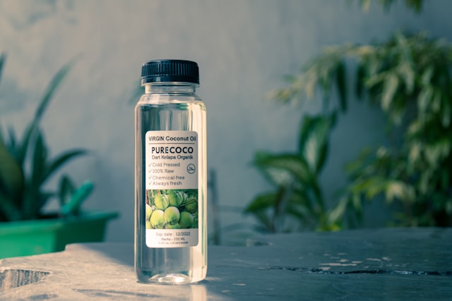 A clear bottle of coconut oil placed on a gray surface with green plants
