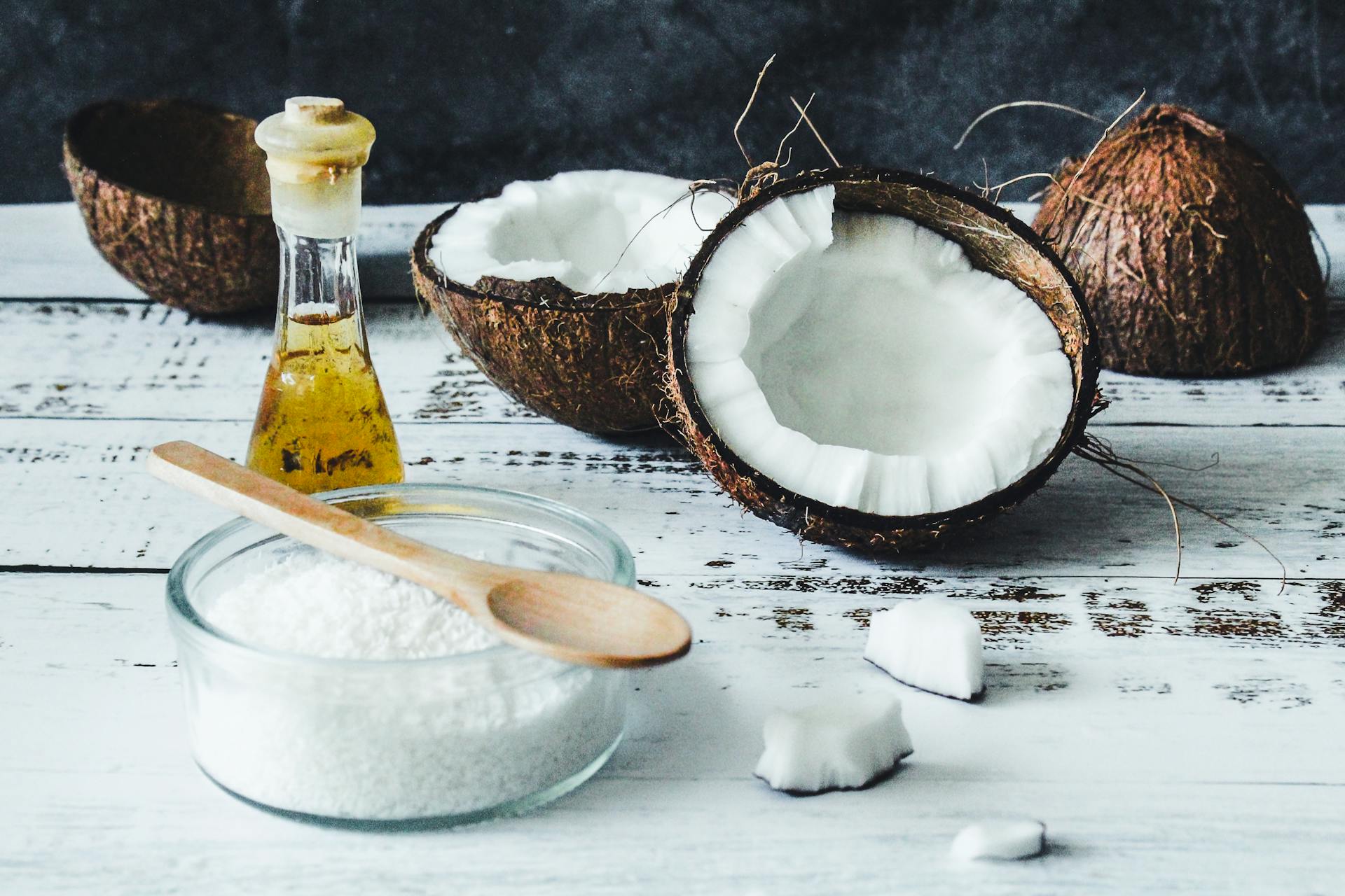 Halved coconuts, a small glass jar of coconut oil, and a bowl of grated coconut with a wooden spoon on a white rustic wooden table