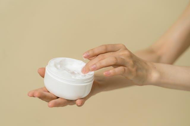 A person dipping fingers into a white jar of creamy coconut-based moisturizer